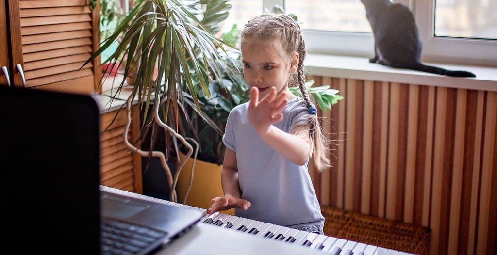 Young girl waving at a laptop screen during online piano lessons for kids