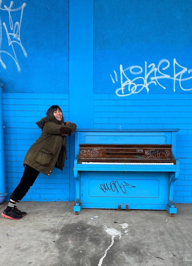 Piano teacher Michelle smiling and leaning on a vintage blue acoustic piano. The piano is sat against a bright blue brick wall.