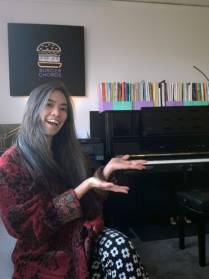 Piano teacher Michelle seated in her piano studio, gesturing her hands towards a black Yamaha upright piano.